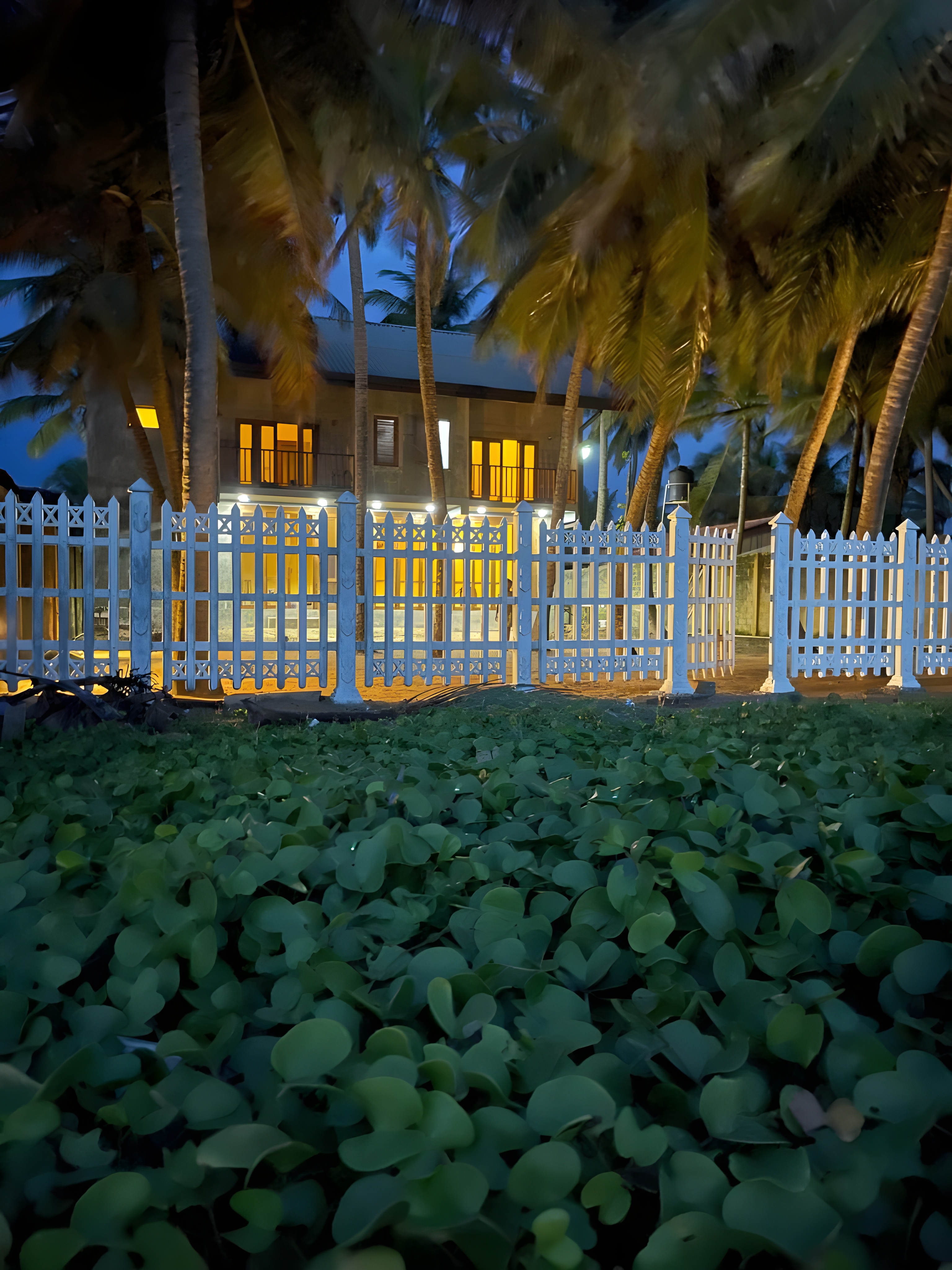 CocoBaru villa at night behind a white beachfront fence and palm trees